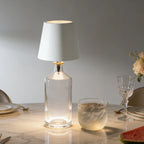 Clear glass table lamp with white shade on a marble surface with dining items.