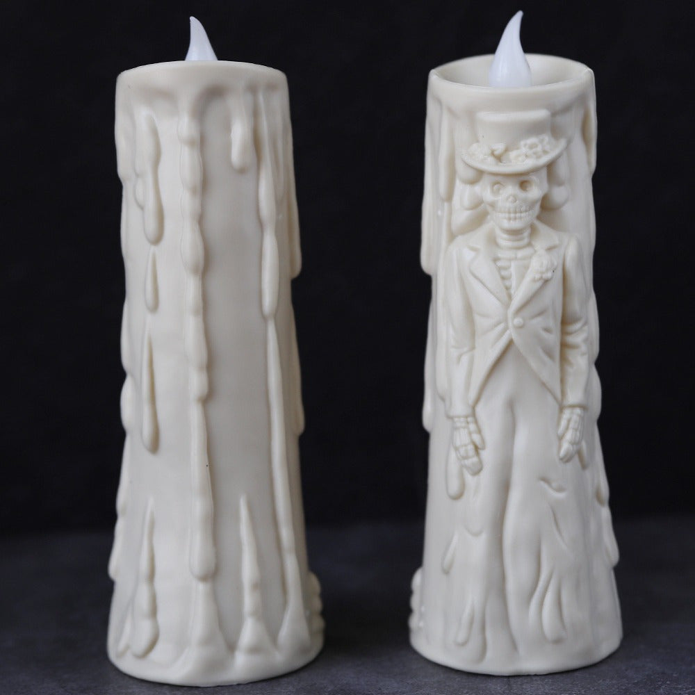 Two white wax candles with flame tips, shown against a dark background. One candle has a dripping wax design, and the other features an embossed skeleton figure in formal attire.