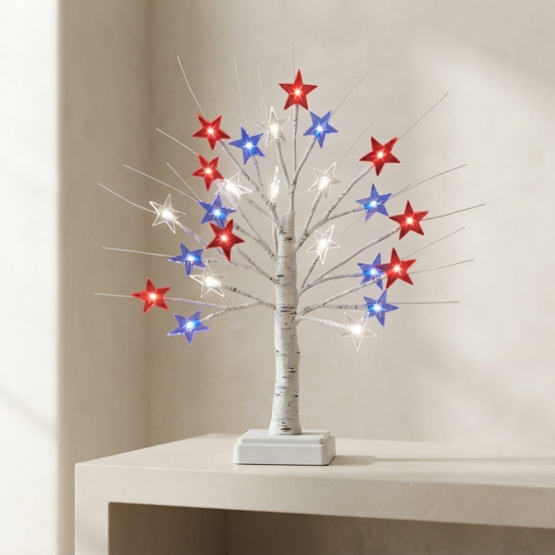A decorative tabletop tree with a white faux-birch trunk and thin branches, lit with small white bulbs and adorned with red, blue, and white star-shaped lights, displayed on a light beige surface against a neutral wall.