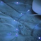 Close-up of clear string fairy lights glowing with blue light, draped over a wet surface with water droplets.