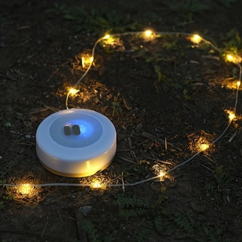 A small round white solar LED string light with a glowing blue indicator light on the top, placed on dark soil outdoors with warm yellow lights winding around it.