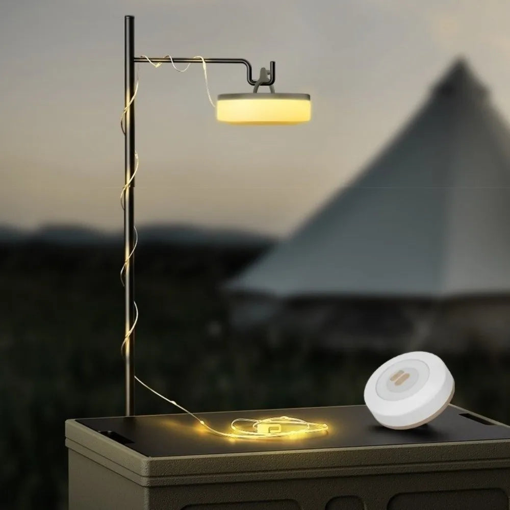 A black metal solar-powered LED light with a glowing yellow lamp and decorative string lights, shown on a dark table with a white charging dock in a blurred outdoor camping setting at dusk.