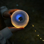 A person holds a round, translucent solar LED light that glows with a warm yellow-orange light from the bottom and a cool blue light from the top, with a string of warm fairy lights visible in the dark background.