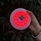 A hand holding a round, light-up LED beauty device with a glowing red interior and a pale pink outer rim, set against a dark background of green leaves.