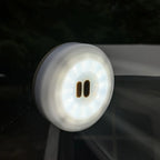 A round white LED window light with a magnetic suction cup base, shown illuminated on a dark glass surface.