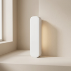 A minimalist white rectangular wall light with rounded edges, standing on a beige windowsill and casting a warm yellow glow against a beige wall.