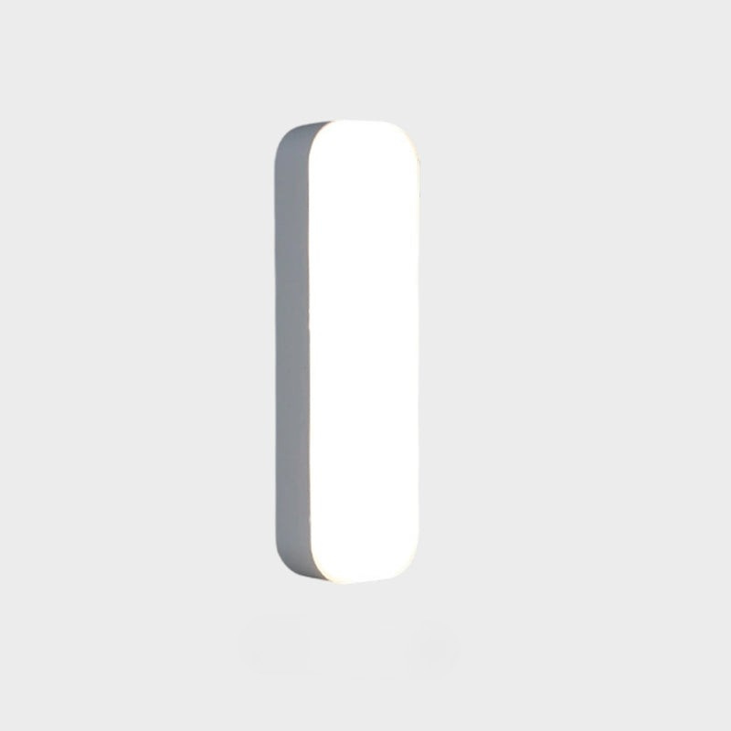 A modern, minimalist vertical rectangular wall light with rounded corners, featuring a glowing white front face and a dark gray side profile, set against a solid light gray background.