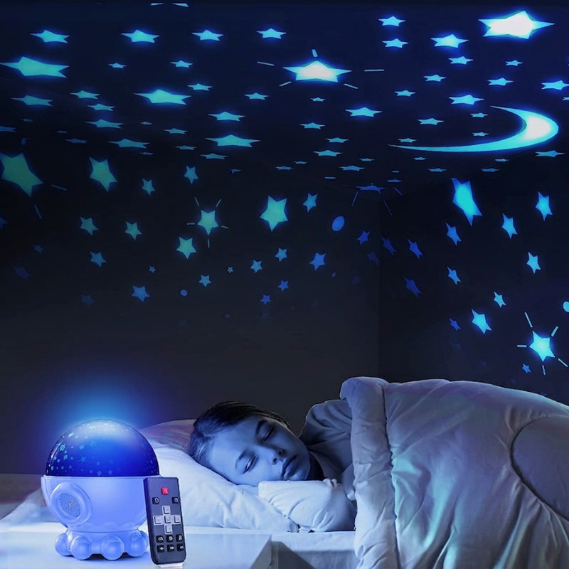 A child's bedroom scene at night with a person sleeping under a blue star and moon projector lamp casting light across the ceiling and walls.