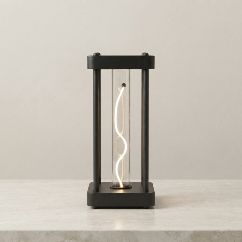 A modern black metal lantern-style table lamp with a clear glass body and a glowing spiral filament, set on a light stone surface against a neutral beige wall.