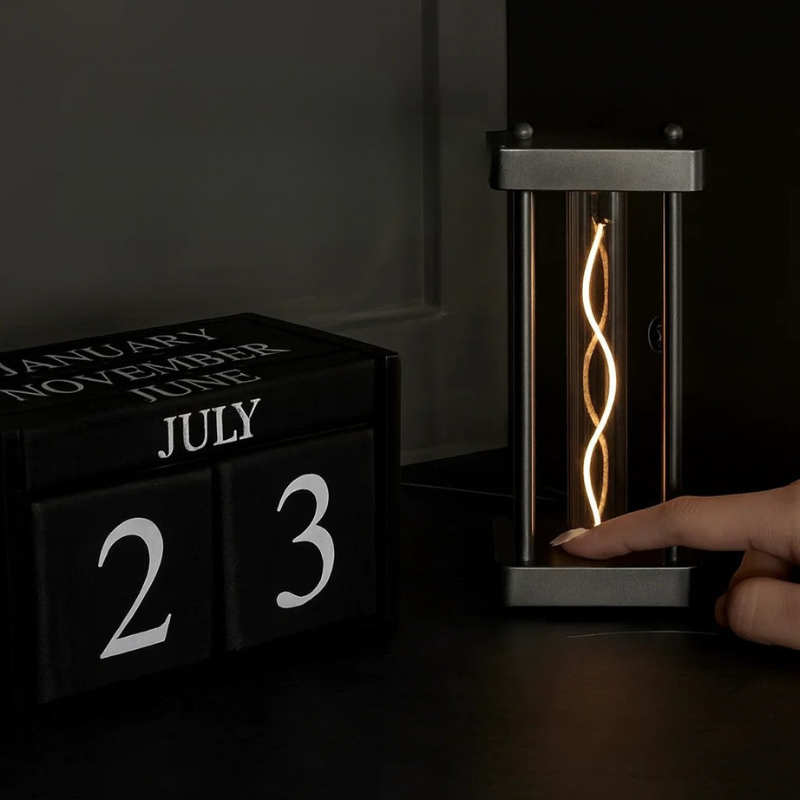 A black block calendar displaying July 23 alongside a modern cylindrical LED filament lamp with a glowing spiral light, shown on a dark tabletop with a hand touching the lamp.