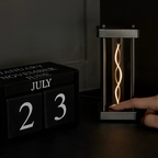 A black block calendar displaying July 23 alongside a modern cylindrical LED filament lamp with a glowing spiral light, shown on a dark tabletop with a hand touching the lamp.