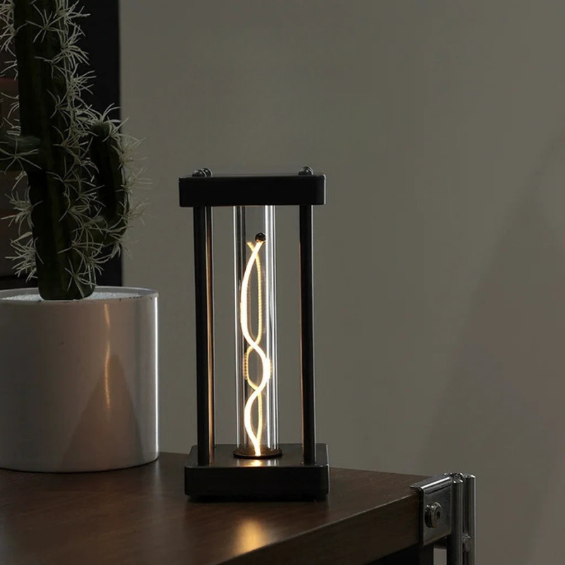 A modern black metal lantern-style table lamp with a glowing spiral filament bulb inside a clear glass or acrylic tube, placed on a wooden surface next to a potted cactus.