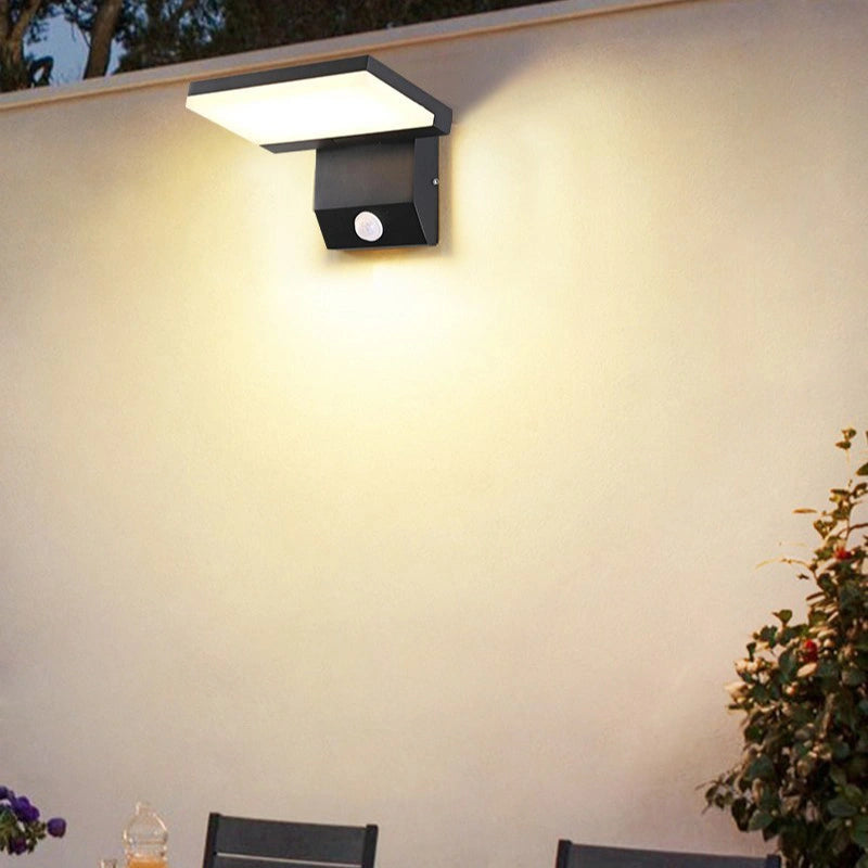 Outdoor wall light fixture on a white wall with a plant and chair in the foreground.