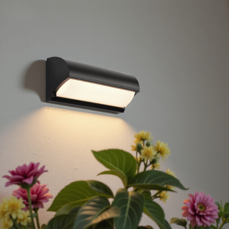 Black outdoor wall light fixture on a gray wall with plants and flowers in the foreground.