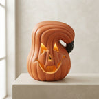 An orange jack-o'-lantern-shaped decorative lamp with carved facial features and a glowing warm light inside, displayed on a light stone pedestal against a soft beige wall.