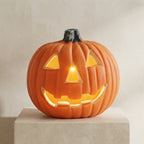 An orange jack-o'-lantern Halloween decoration with a carved face, glowing with warm yellow light from within, displayed on a light beige pedestal against a neutral beige background.