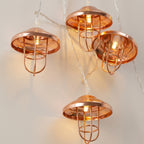 Mushroom-shaped string lights with copper-colored metal caps and wire cages, glowing with warm white bulbs on a light beige wall.