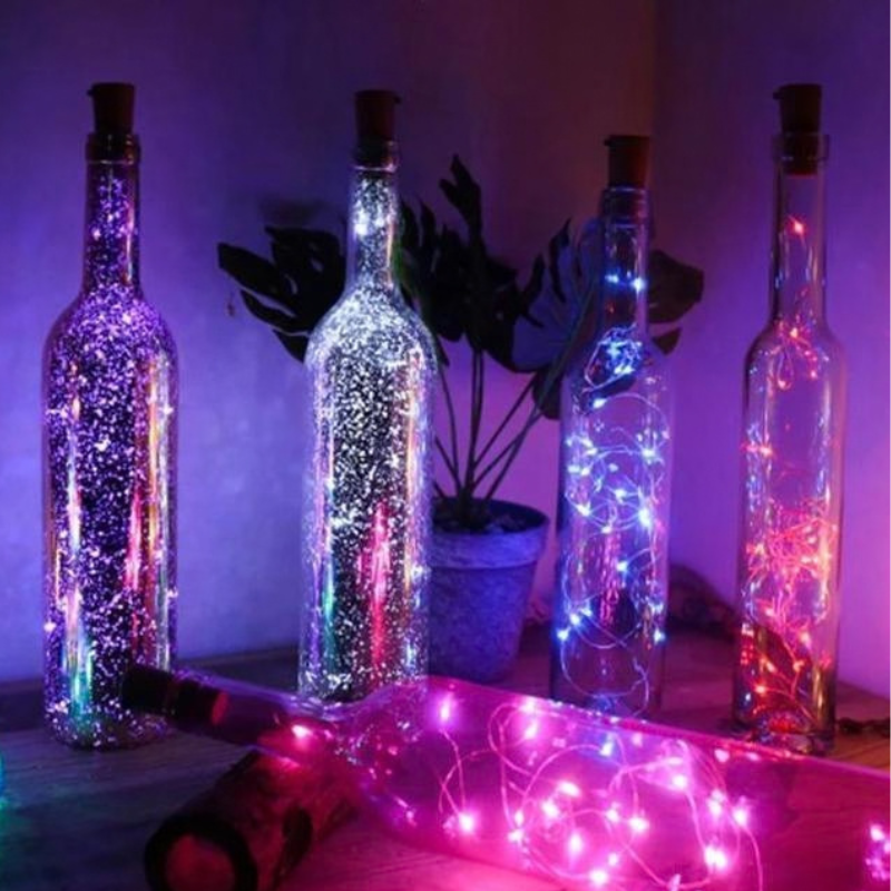 Four clear glass bottles with cork stoppers, each filled with glowing fairy string lights in different colors including purple, white, blue, and pink. A fifth bottle lies in the foreground on its side, showing bright pink lights inside.