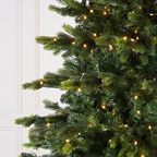 Close-up of a green artificial Christmas tree with warm white string lights, shown against a white paneled wall.