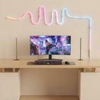 A modern desk setup featuring a curved monitor displaying a neon-lit city scene, flanked by two slim vertical speakers, with a keyboard and mouse on a desk below. Above the desk, a wavy neon light sign glows in pink, blue, and white against a beige wall.