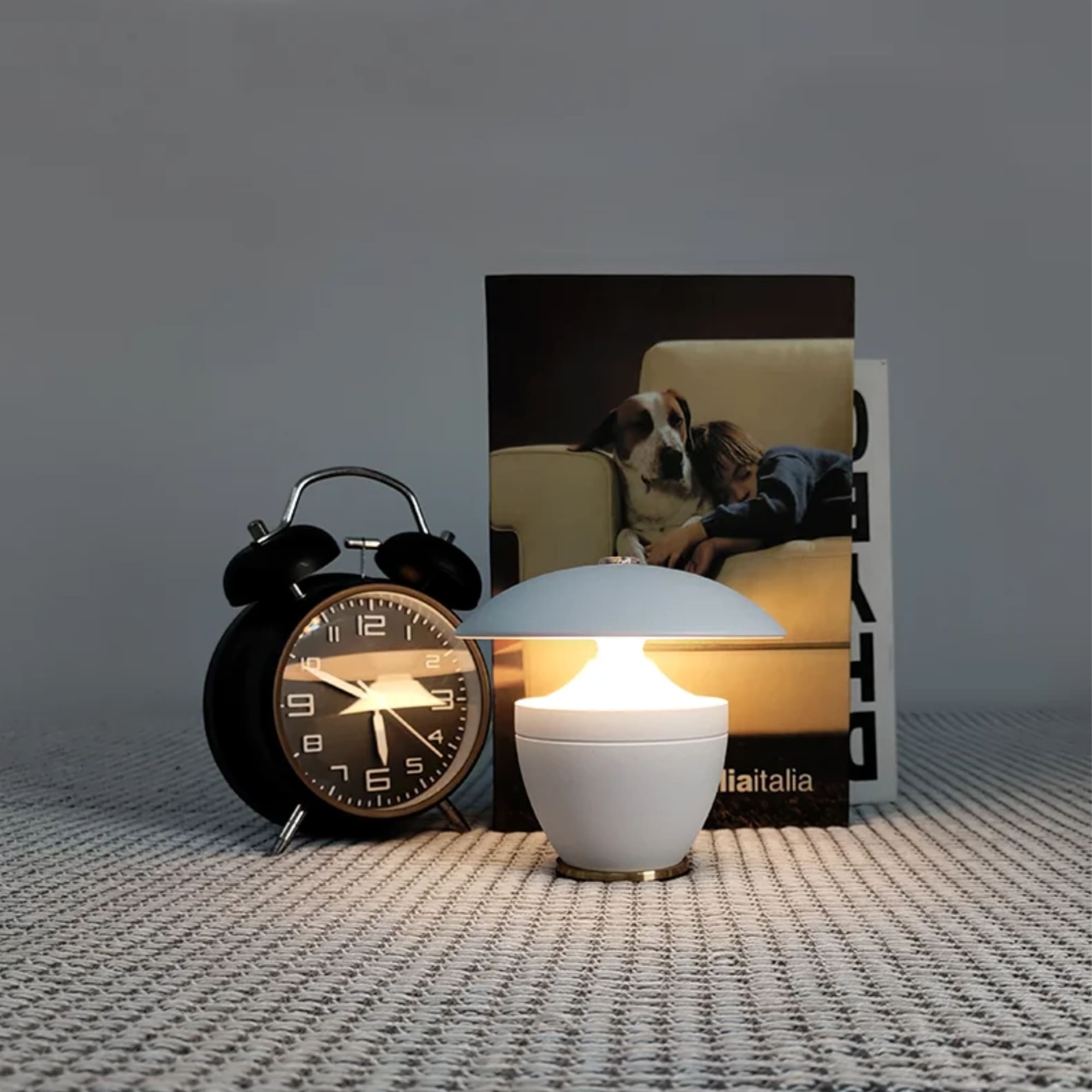 Black alarm clock and white lamp on a textured surface with a magazine featuring a dog on a couch in the background.