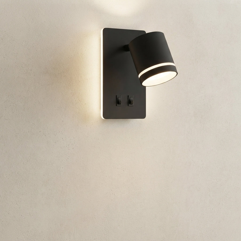 A modern black wall sconce with a cylindrical spotlight and a rectangular backplate featuring two switches, mounted on a textured beige wall and casting a warm light.
