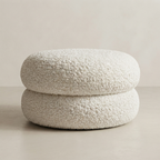 A round, two-tiered pouf or ottoman upholstered in a textured, boucle-like cream fabric, set against a minimalist beige background.