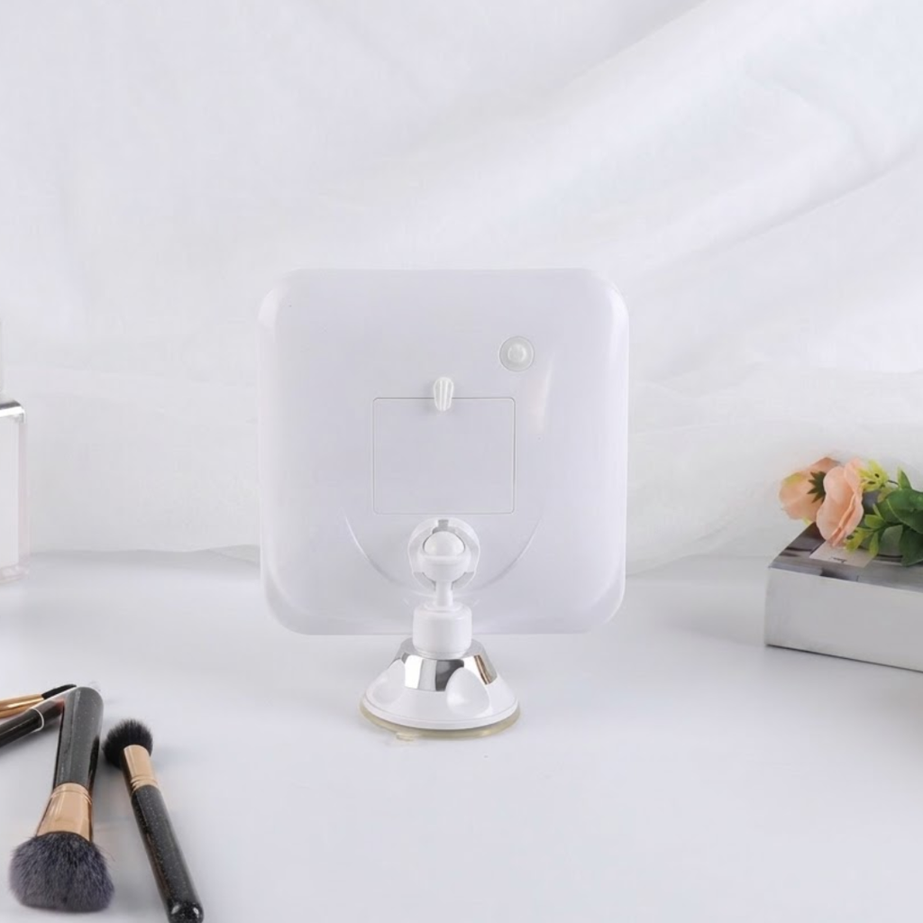 A white square wall-mounted mirror with a clear suction cup base, shown on a white tabletop with makeup brushes and a small flower arrangement in the background.