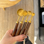 Set of four gold measuring spoons with wooden handles held by a hand against a neutral background.
