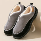 A pair of gray slip-on winter slippers with a black sole, cream sherpa lining, and decorative silver stitching, displayed on a beige background.
