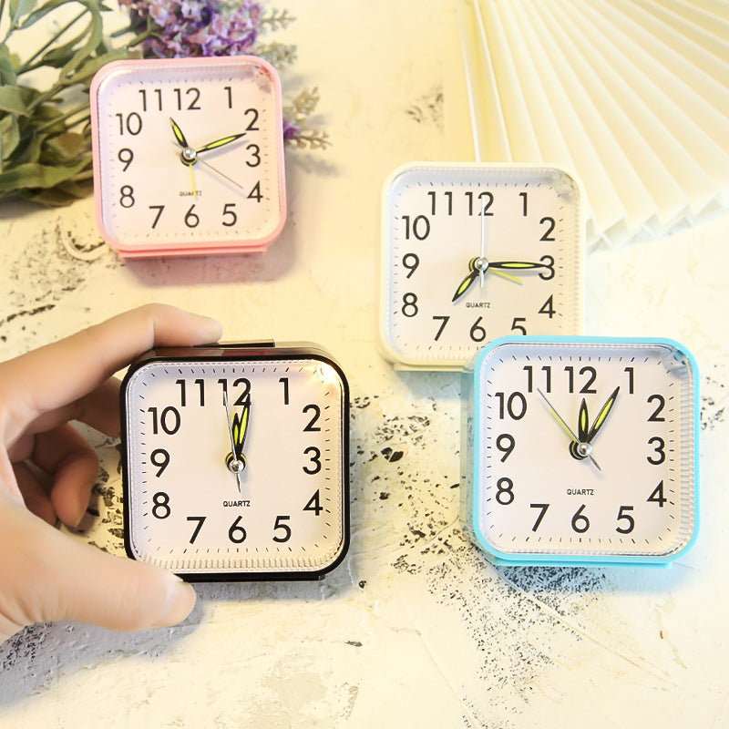 Four small square clocks in different colors on a textured surface with a hand pointing to one of them.