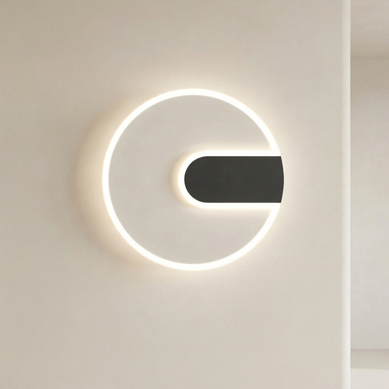 A modern minimalist wall sconce featuring a glowing white circular ring with a black horizontal bar extending from the center, mounted on a warm off-white wall.