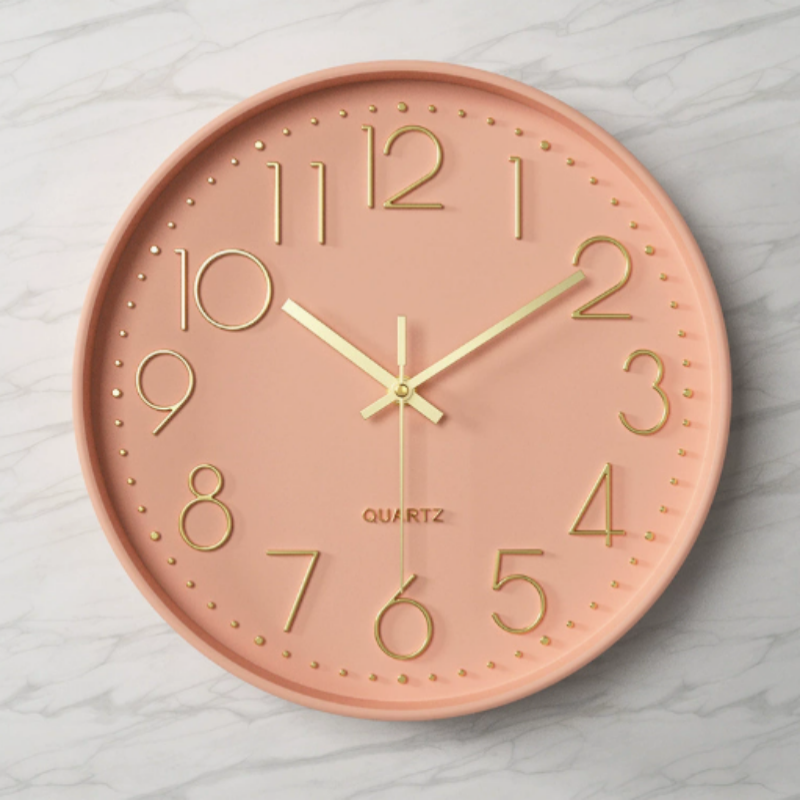 A round wall clock with a dusty pink face and gold-tone numerals, hands, and markers, mounted on a light gray marble-patterned wall.