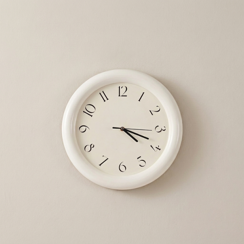A minimalist round wall clock with a white frame and a white dial, featuring black Arabic numerals and black hands, mounted on a light beige wall.
