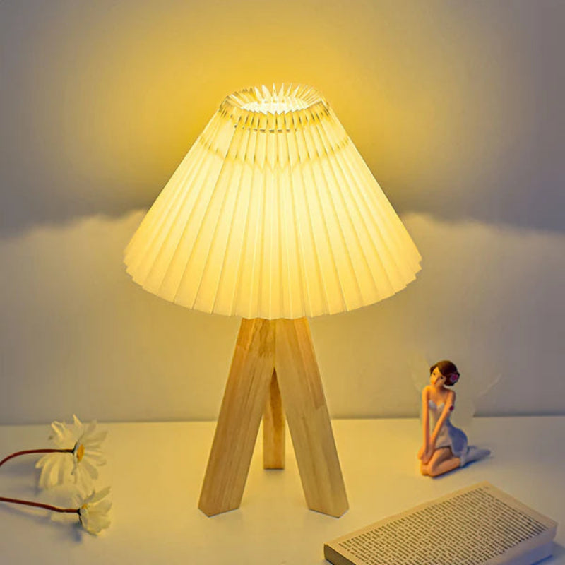 Wooden table lamp with pleated shade on a white surface, surrounded by decorative items.