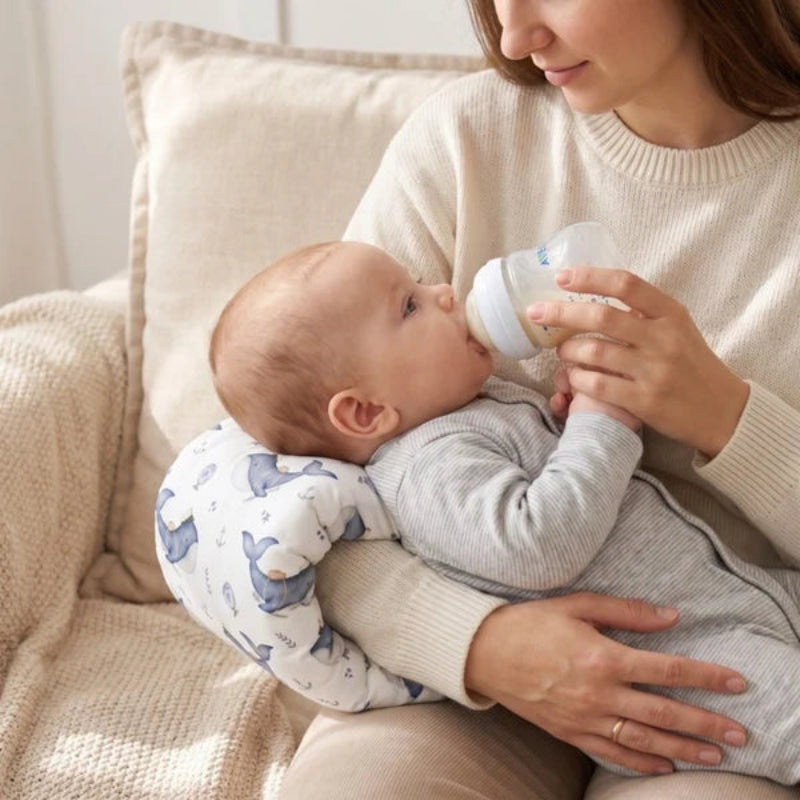 A baby wearing a gray long-sleeve romper rests on a woman's lap on a beige couch, drinking from a white bottle while supported by a blue and white patterned baby support pillow.