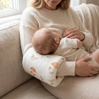 A woman in a cream sweater holds a baby on her lap while breastfeeding. The baby wears a matching cream outfit and rests on a patterned nursing pillow with orange mushrooms and green leaves, set on a beige sofa near a window.