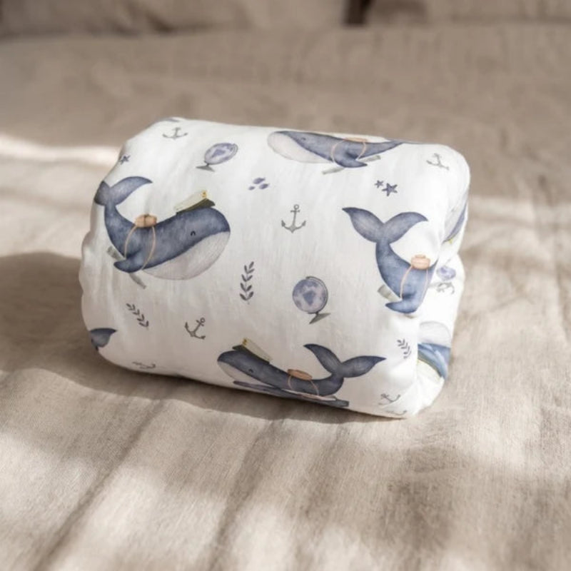 A rolled-up white fabric item, likely a baby swaddle or blanket, printed with a navy blue whale and small anchor motifs, resting on a beige linen surface with soft natural shadows.