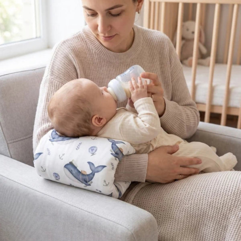 A woman sitting on a light gray armchair feeds a baby from a bottle while the baby rests on a whale-print baby support pillow.