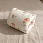 A rolled-up cream-colored fabric item, likely a pillow or cushion cover, printed with a watercolor-style pattern of orange-brown mushrooms and small green leaves, resting on a beige linen surface.