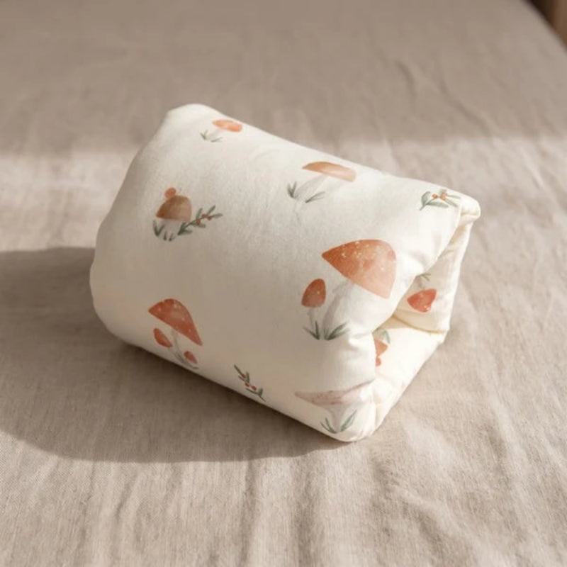A rolled-up cream-colored fabric item, likely a pillow or cushion cover, printed with a watercolor-style pattern of orange-brown mushrooms and small green leaves, resting on a beige linen surface.