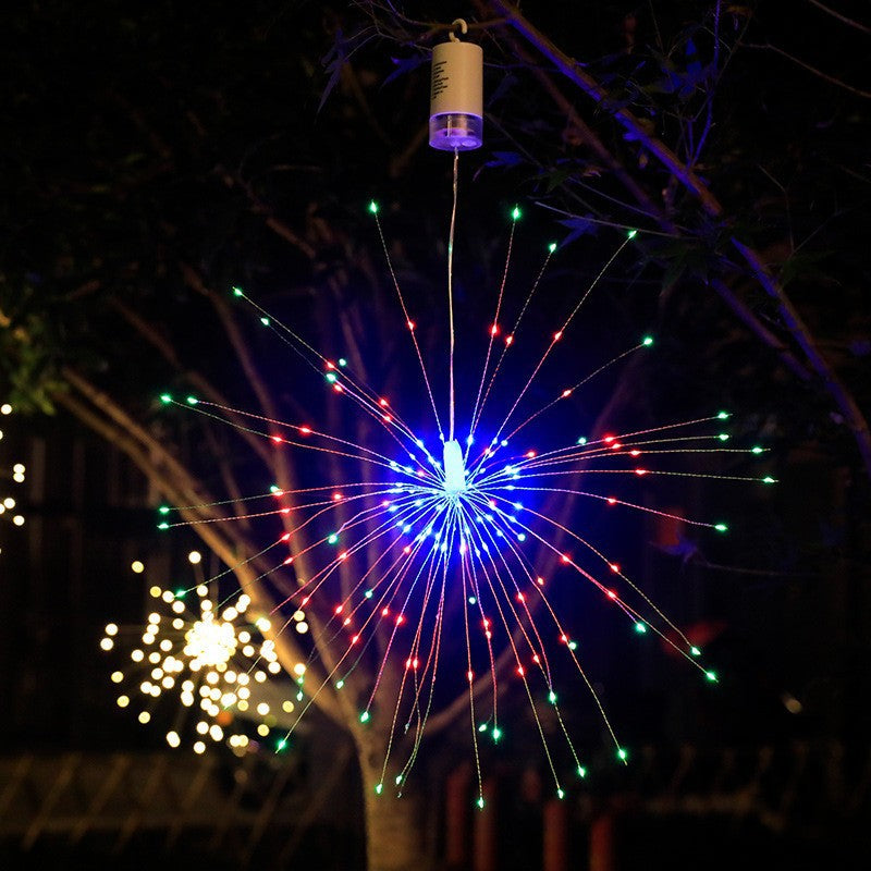 A hanging starburst LED light fixture with multiple thin strands emitting red, green, and blue light rays, suspended from a cylindrical top piece against a dark night scene with trees and blurred lights.