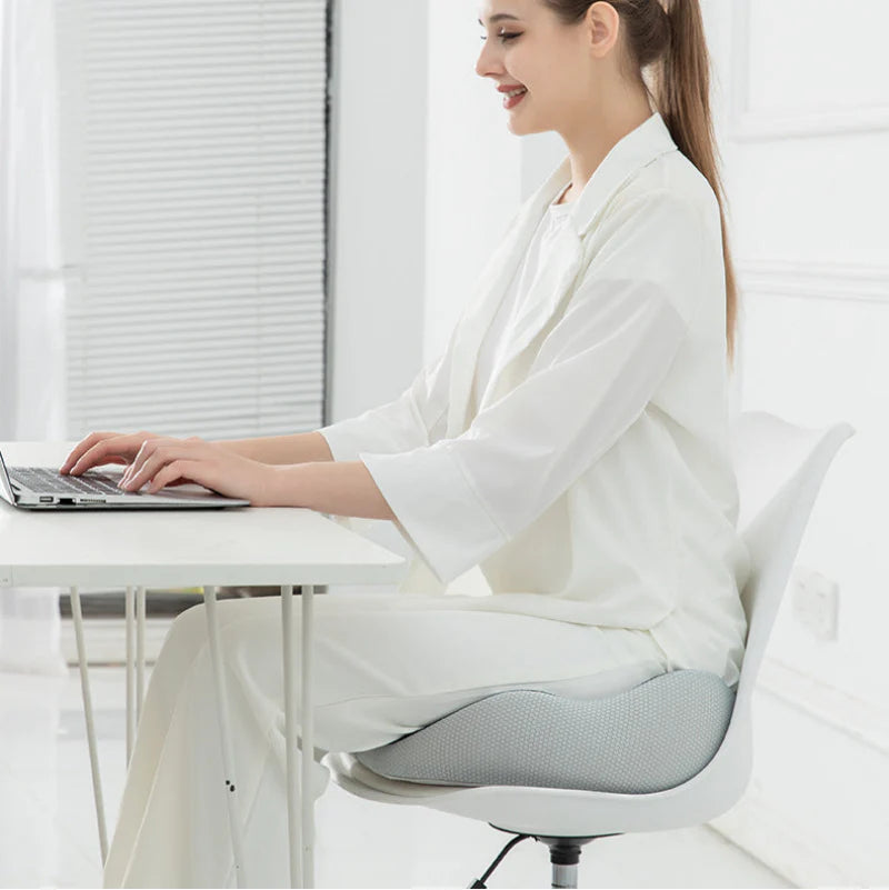 Woman sitting on a white chair with a gray cushion, using a laptop in a bright room.