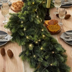 A green pine garland with warm white string lights draped across a wooden dining table, surrounded by plates, wine glasses, bread, and candles in a cozy holiday setting.