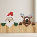 A festive holiday wall decoration set featuring a smiling Santa Claus head with a red hat, white beard, and green mittens on the left, and a brown reindeer head with gray antlers on the right, both peeking over a wooden panel against a light beige wall.