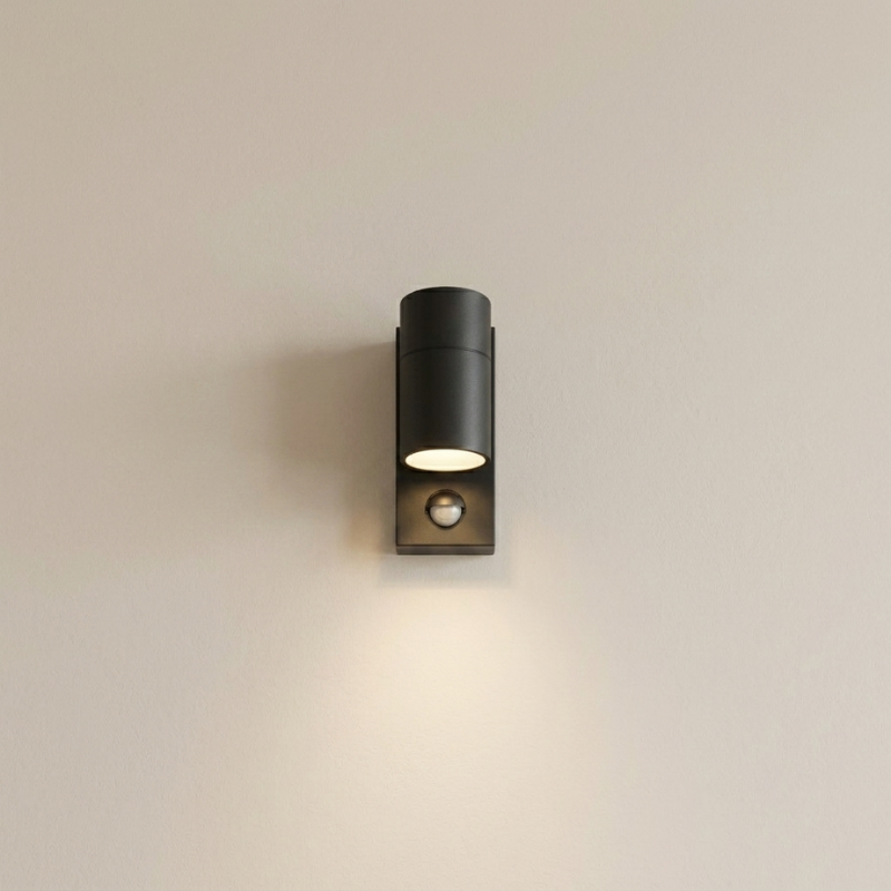 A modern black cylindrical outdoor wall sconce with a built-in motion sensor, mounted on a plain beige wall and casting a warm light downward.