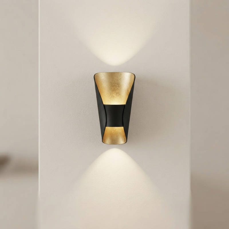 A modern wall-mounted sconce with a black outer frame and textured gold inner panels, shown lit against a warm off-white wall.