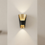 A modern wall-mounted sconce with a black outer frame and textured gold inner panels, shown lit against a warm off-white wall.