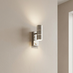 A modern cylindrical wall sconce with a brushed silver finish and integrated warm white LED lighting, mounted on a beige textured wall.