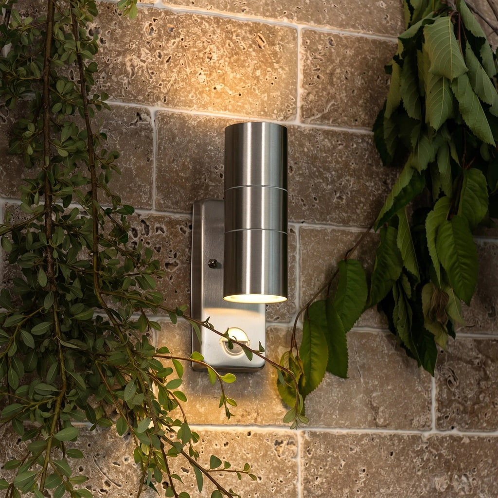 A modern cylindrical outdoor wall sconce with a brushed silver metal finish, mounted on a textured stone wall and illuminated to cast a warm glow, framed by green foliage.